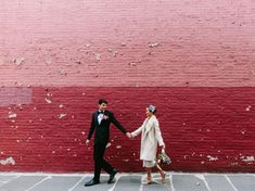 a man and woman holding hands walking in front of a red brick wall with their arms around each other