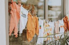 several baby ones hanging on a clothes line in front of a store window with an orange potted plant next to it