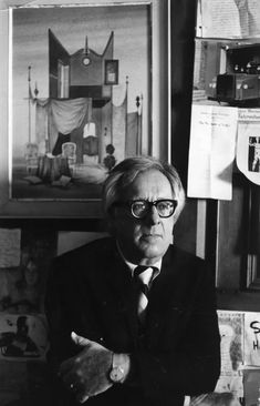 black and white photograph of a man in glasses sitting at a desk with papers on the wall behind him