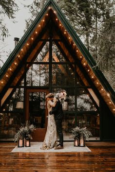 a bride and groom standing in front of a cabin with lights on the roof, surrounded by greenery