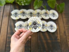 a hand holding up a bee sticker on top of a wooden table next to green plants