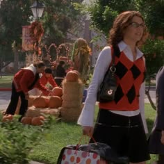 a woman in a red and black argyle sweater carries her luggage down the street with other people dressed up as pumpkins