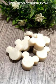 some white candles sitting on top of a wooden table next to green leaves and flowers