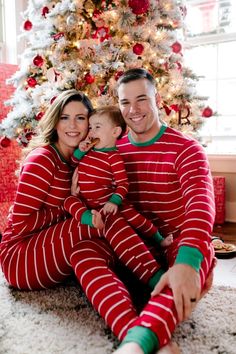 a man and woman in matching pajamas are sitting next to a christmas tree while holding their child