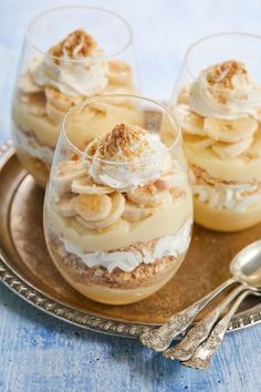 three desserts with bananas and whipped cream on a silver platter next to two spoons