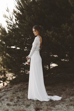 a woman in a long white dress standing next to some trees and looking off into the distance