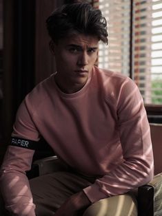 a young man in a pink sweater sitting on a chair next to a window with blinds