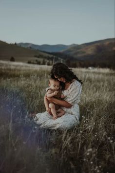 a woman holding a baby sitting on top of a grass covered field with mountains in the background