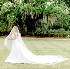 a woman in a wedding dress and veil walking through the grass with trees behind her