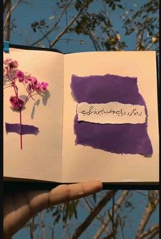 a hand holding an open book with purple and white paint on it, in front of some trees