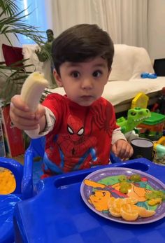 a young boy holding a banana in front of a plate of fruit on a blue table