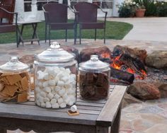 three glass jars filled with marshmallows sit on a table in front of a fire pit