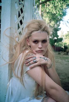 a woman with tattoos on her face and arm sitting in front of a white fence