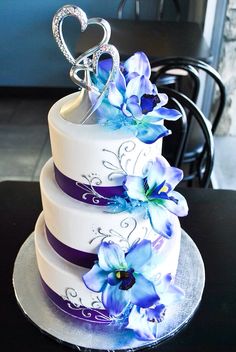 a three tiered wedding cake with blue flowers on the top and purple ribbon around the bottom