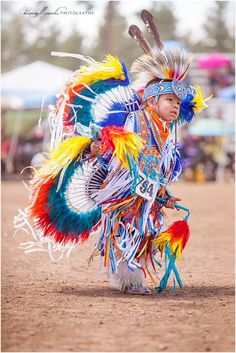 a young child dressed in colorful feathers and headdress, dancing on dirt ground