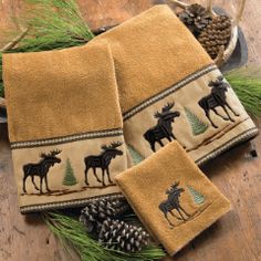 three hand towels with moose and pine cones on them sitting on a wooden floor next to pine cones