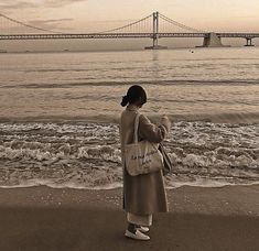 a woman standing on top of a sandy beach next to the ocean with a bridge in the background