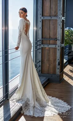 a woman standing in front of a window wearing a long sleeved wedding dress with an open back