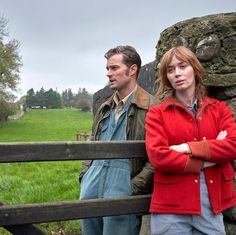 two people standing next to each other in front of a stone wall and wooden fence