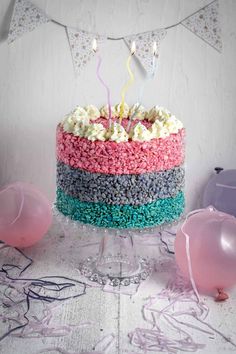 a multicolored cake sitting on top of a table next to balloons and confetti