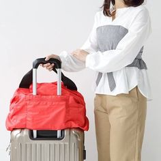 A woman standing with a grey suitcase with a large red bag through the handle she holds. Functional Foldable Travel Accessories For On-the-go, Practical Large Capacity Travel Accessories For On-the-go, Multifunctional Duffle Bag For Daily Use, Red Large Capacity Gym Bag For Travel, Large Capacity Red Gym Bag For Travel, Practical Red Travel Bag, Versatile Gym Bag With Luggage Sleeve For Trips, Multifunctional Travel Duffle Bag, Functional Gym Bag With Pockets For Weekend Trips