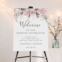 a welcome sign with pink flowers on it in front of a white wall and string lights