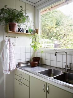 a kitchen sink under a window with potted plants on the ledge and hanging towels