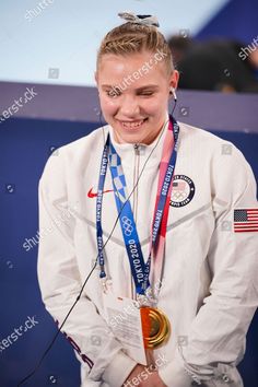 a woman in white jacket holding two medals and smiling at the camera with her hands behind her back