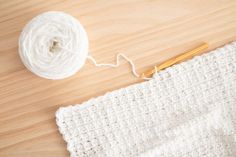 a white crochet blanket next to a ball of yarn and a knitting needle