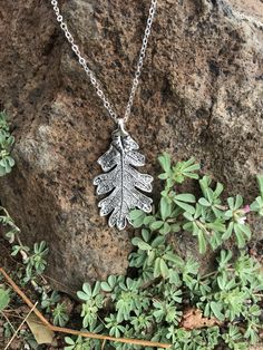 a silver necklace with an oak leaf on it sitting next to some plants and rocks