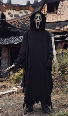 a man in a skeleton mask and black cloak holding a knife standing outside an old house