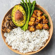 a wooden bowl filled with rice, asparagus and tofu next to sliced avocado