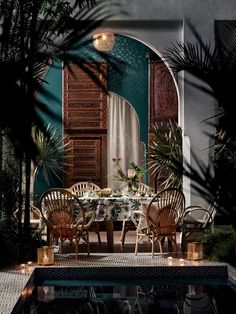 an outdoor dining area with wooden chairs and table surrounded by greenery in front of a doorway