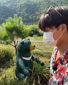 a woman wearing a face mask standing next to a stuffed bear in the grass with trees and mountains behind her