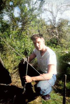 a man squatting down next to a black horse in the grass with trees and bushes behind him