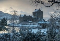 an old castle with snow on the ground and trees around it in front of a body of water