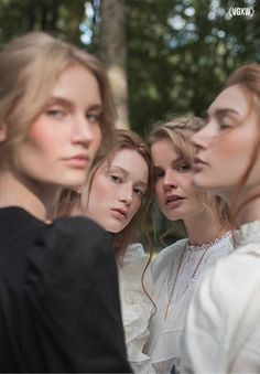 three women are standing in front of trees and one is looking at the other woman's face