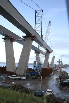 the bridge is being built over the water and cars are parked on the road below it