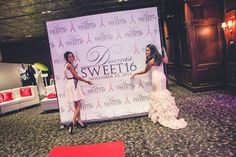 two women standing in front of a sign at a fashion show with red carpet and white walls