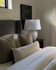 a bed with white sheets and pillows next to a lamp on a table in front of a window