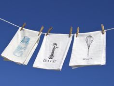 three tea towels hanging on a clothes line with kitchen utensils and whisks