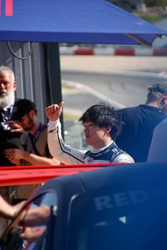 a man waves to the crowd as he stands in front of a blue car with his hand up