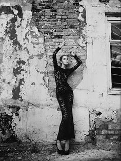 black and white photograph of woman in long dress leaning against an old brick building with peeling paint