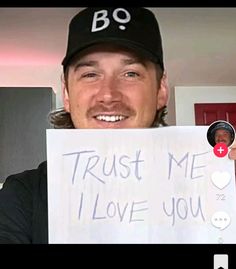 a man holding up a sign that says trust me i love you and he is wearing a baseball cap