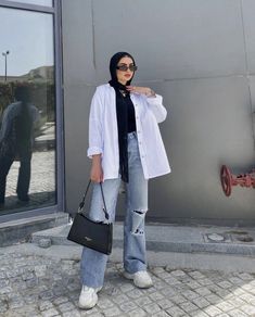 a woman standing in front of a building holding a black purse and wearing jeans with ripped knees