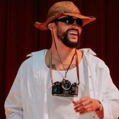 a man wearing a hat and holding a camera in front of a red curtain with his hands out