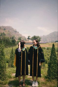 two women in graduation gowns standing next to each other on the grass with trees behind them