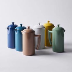 four different colored coffee pots and one teapot on a white surface with the lid off