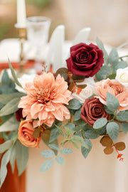 an arrangement of flowers and greenery on a white table cloth with candles in the background