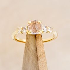 a gold ring with an oval morganite surrounded by three small white diamonds on a wooden stand